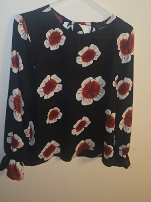 Svart blus med stora blommor - Svart blus med stora vita och röda blommor. Blusen har långärmade ärmar och en liten knytning i nacken. Tunt och lätt material som ger ett luftigt intryck. Perfekt för dig som gillar ett färgstarkt mönster.