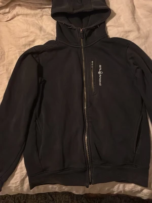 Svart hoodie från Sail Racing - Svart hoodie från Sail Racing i storlek S med dragkedja framtill och en bröstficka med dragkedja. Snyggt Sail Racing-tryck på bröstet och klassisk huva. Perfekt för en sportig och avslappnad stil.