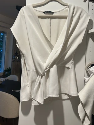 Vit omlottblus från Zara - Stilren vit omlottblus från Zara med knytning i midjan och djup v-ringning. Blusen har korta ärmar och är tillverkad i ett mjukt, lätt material som ger ett snyggt fall. Perfekt att matcha med jeans eller kjol för en trendig look.