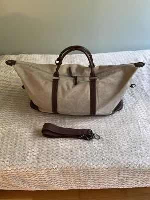 Beige och brun weekendbag - Snygg och rymlig weekendbag i beige canvas med bruna detaljer i konstläder. Väskan har två handtag, avtagbar axelrem och dragkedja längs mitten. Perfekt för resor när du vill ha både stil och funktion. Unik form och robust material gör den till ett riktigt statement.