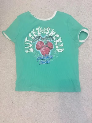 Grön t-shirt med tryck och vita kanter - Snygg grön t-shirt med vita kanter vid hals och ärmar. Framsidan har ett färgglatt tryck med hallon och texten 'Out of this world positive mind'. Perfekt för dig som gillar färg och coola prints.