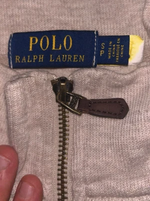 Beige halvzip från Polo Ralph Lauren - Snygg beige halvzip-tröja från Polo Ralph Lauren med klassisk broderad logga på bröstet. Tröjan har en metallblixtlås med läderdetalj och är tillverkad i mjukt material, perfekt för en avslappnad men stilren look.