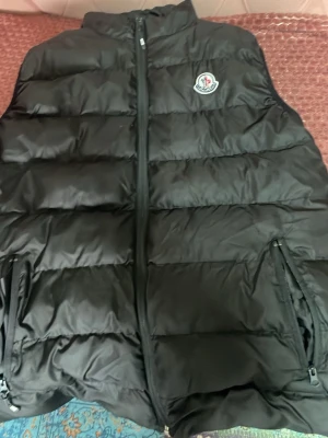 Svart dunväst från Moncler - Snygg svart dunväst från Moncler med klassisk quiltad design och dragkedja framtill. Västen har två fickor med dragkedja och Monclers ikoniska logga på bröstet. Perfekt för lager-på-lager och håller dig varm under kyliga dagar.