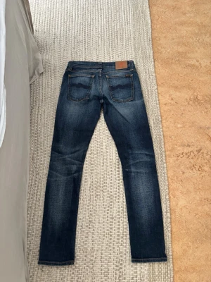 Nudie jeans - Snygga mörkblå jeans med raka ben och klassiska kontrastsömmar. Jeansen har två bakfickor med vågig detalj och en brun patch i midjan. Materialet är denim med lätt tvättad effekt för en avslappnad look.