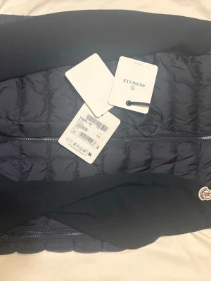  Moncler jacka - Snygg mörkblå dunjacka från Moncler med quiltad front och stickade ärmar. Jackan har dragkedja framtill, hög krage och Moncler-logga på ärmen. Perfekt för kalla vinterdagar och riktigt stilren design.