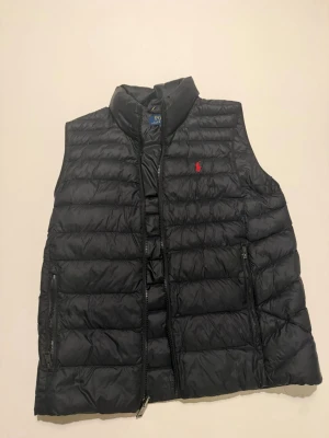 Svart dunväst från Polo Ralph Lauren - Snygg svart dunväst från Polo Ralph Lauren med klassisk dragskedja och två par sidofickor. Ett litet broderat rött Polo märke.