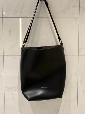 Svart axelväska från Claudia Canova - Stilren svart axelväska i slätt konstläder från Claudia Canova. Väskan har en justerbar axelrem och minimalistisk design med diskret logga framtill. Perfekt för dig som gillar enkel och modern stil, helt ny skick 🖤