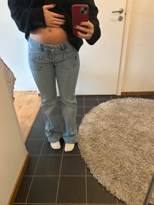 Ljusblå bootcut jeans med fickor - Säljer ett par ljusblå jeans med bootcut passform och höga fickor fram. Jeansen har synliga knappar och är tillverkade i klassisk denim. Perfekta för en avslappnad och trendig look.