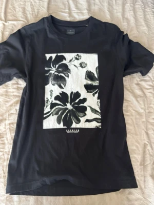 Svart blommig t-shirt Jack & Jones - Snygg svart t-shirt från Jack & Jones med stort blommigt tryck i vitt och svart på bröstet. Klassisk passform och rund halsringning. Perfekt till jeans eller shorts för en avslappnad look.