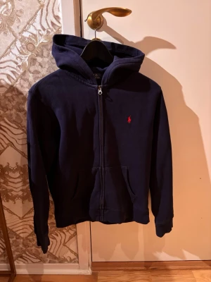 Mörkblå Polo Raphael Lauren Kofta - Snygg mörkblå kofta med dragkedja och huva. Klassisk design med fickor framtill och en liten röd logga broderad på bröstet. Perfekt för en avslappnad stil och enkel att matcha med jeans eller joggers. OBS STORLEKEN ÄR I BARN (14-16år) (ish 160-170cm)