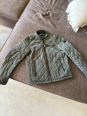 Olivgrön quiltad jacka från Replay - Snygg olivgrön quiltad jacka från Replay med dragkedja framtill och två snedställda fickor. Jackan har lång ärm, ståkrage och diskret Replay-logga på ärmen. Perfekt för dig som vill ha en stilren och bekväm jacka.