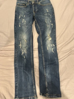 Blå slitna raka jeans - Säljer ett par blå jeans med raka ben och slitna detaljer framtill. Jeansen har klassisk femficksmodell och kontrastsömmar. Materialet är denim i bomull och de har en cool, avslappnad look med distressed effekter.