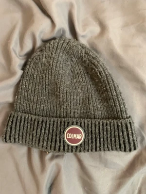 Grå ribbstickad mössa från Colmar - Säljer en grå ribbstickad mössa från Colmar Originals. Mössan har uppvikt kant och en rund Colmar-logga framtill. Tillverkad i mjukt och varmt material som passar perfekt för kyliga dagar. Enkel och stilren design som funkar till det mesta.