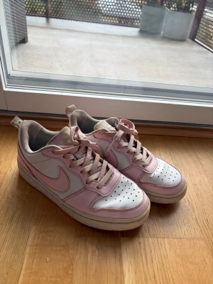 Vita och rosa Nike sneakers - Säljer ett par vita och ljusrosa Nike sneakers med klassisk swoosh på sidan. Skorna har låga skaft, perforerad tåbox och matchande rosa snören. Tillverkade i läder med gummisula. Perfekta för dig som gillar en clean och sportig look.                                     (Skosnören inte tvättade) 