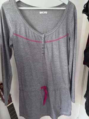 Grå långärmad klänning Tom Tailor - Grå klänning från Tom Tailor med lång ärm, knappar framtill, rosa dekorlinje över bröstet och rosa knytband i midjan. Mjuk och enkel modell med rundad halsringning, perfekt för en chill och snygg vardagsstil. Vet inte storlek men gissar på S/M!