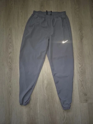 Grått Nike träningsset - Säljer ett grått träningsset från Nike med matchande byxor och jacka. Byxorna har resår i midjan och vid bensluten samt fickor på sidorna. Jackan har huva och reflexdetaljer på ärmarna. Perfekt för löpning eller gym. Materialet är lätt och funktionellt.