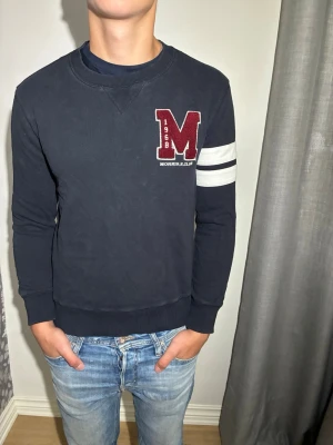Mörkblå Morris sweatshirt - Snygg mörkblå sweatshirt med ett stort rött M-tryck och vita detaljer på bröstet. Två vita ränder på ena ärmen ger en sportig look. Rund halsringning och ribbade muddar. Perfekt till jeans för en avslappnad stil. Han på bilden är 180 och väger 80kg