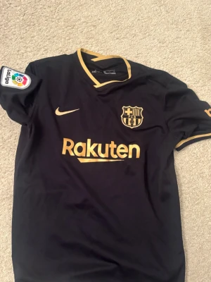 Svart FC Barcelona matchtröja Nike - Snygg svart FC Barcelona matchtröja från Nike med guldfärgade detaljer och loggor. Tröjan har Rakuten och UNICEF-tryck, La Liga-märke på ärmen och guldfärgade ribbade kanter. Perfekt för dig som älskar fotboll och vill representera Barca. Storlek xs 