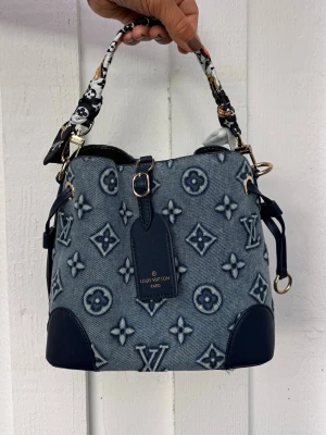 Blå trendig väska i denim Ny, tidlös och klassisk  - Trendig handväska i blått denimtyg med klassiskt monogrammönster och mörkblåa läderdetaljer. Väskan har en kort handtag med svartvitt mönster och guldfärgade metalldetaljer. Perfekt accessoar för dig som gillar lyxig streetstyle.
