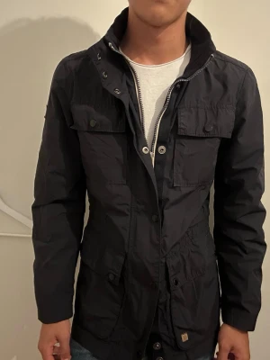 Svart field jacket med hög krage - Stilren svart skepparkavaj med hög krage och flera fickor framtill. Jackan har knappar och dragkedja samt en patch på ärmen. Materialet är lätt och passar perfekt för lager-på-lager. Snygg och modern design som funkar till jeans.