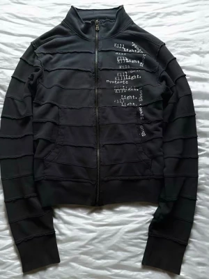 Hysteric glamour zip up - Säljer en svart hoodie med dragkedja och hög krage. Framsidan har vita texttryck med ord som 'Killlight' och 'Shoes'. Hoodien har råa sömmar i horisontella linjer över hela plagget för en edgy look. Materialet känns mjukt och stretchigt, perfekt för en avslappnad stil.