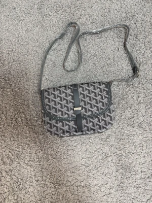 Grå axelväska  - Snygg grå axelväska med klassiskt mönster och justerbar axelrem. Väskan har ett lock med magnetlås och detaljer i läder. Perfekt för dig som vill ha en unik och trendig accessoar med lyxig känsla.