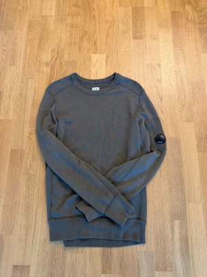 Olivgrön sweatshirt från C.P. Company - Snygg olivgrön sweatshirt från C.P. Company med rund hals och ribbade muddar. Tröjan har en ikonisk linsdetalj på ena ärmen och är tillverkad i mjuk bomull. Perfekt för en avslappnad och stilren look.