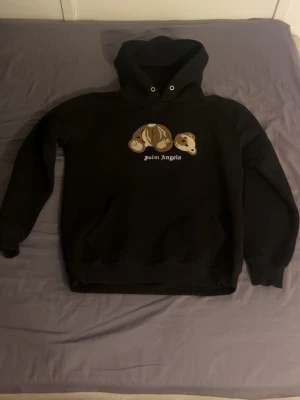 Svart Palm Angels hoodie med björn - Säljer en svart hoodie från Palm Angels med en broderad björn på bröstet och texten 'palm angels' under. Hoodien har huva med dragsko och en stor magficka framtill. Skriv ifall ni undrar något!