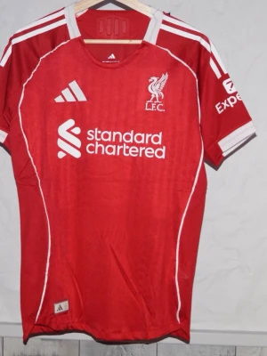 Liverpool röd matchtröja Adidas - Röd Liverpool FC matchtröja från Adidas med vita detaljer och klubbmärke på bröstet. Tröjan har korta ärmar, rund hals och tryckta loggor från Standard Chartered och Expedia. Perfekt för fotbollsträning eller att bära på matchdag. Är du efter en annan storlek är det bara att skriva!