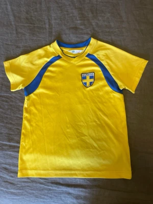 Barn stl. 110/116 Gul Sverige t-shirt från H&M - Gul  t-shirt från H&M med blå detaljer och Sverige-emblem . Fint kvalitet men det finns mörka fläckar på ena armen (syns på bilder)