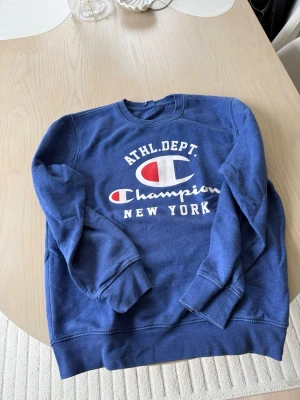 Blå sweatshirt från Champion - Blå sweatshirt från Champion med tryck i vitt och rött på bröstet där det står 'ATHL. DEPT. Champion NEW YORK'. Klassisk rund halsringning och ribbade muddar vid ärmar och nederkant. Perfekt för en sportig och avslappnad stil. Storlek M. Bra skick.