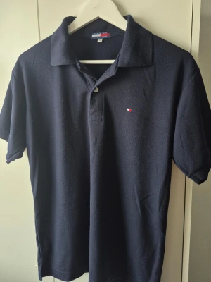 Tommy Hilfiger piké  - Klassisk marinblå pikétröja från Tommy Hilfiger med liten broderad logga på bröstet. Tröjan har krage, korta ärmar och knäppning med två knappar. Perfekt till jeans eller shorts för en clean och stilren look.
