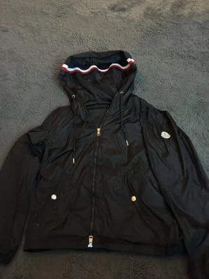 Svart vindjacka från Moncler - Snygg svart vindjacka från Moncler med huva och dragkedja framtill. Jackan har guldfärgade detaljer, två fickor med knappar och Moncler-logga på ärmen. Huvan har en cool kant med röd, vit och blå rand. Perfekt för blåsiga dagar. Passar både L och M