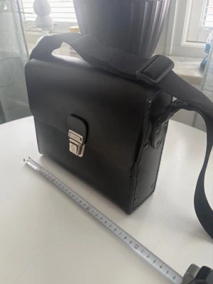 Svart portfölj i läder med axelrem - Snygg svart portfölj i läder med justerbar axelrem och metallspänne framtill. Insidan har flera nyckelringar och fack för smart organisering. Perfekt för dig som vill ha koll på nycklar och småprylar. Klassisk och stilren design.