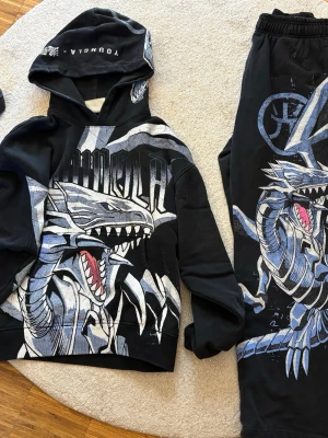 Svart Yu-Gi-Oh! set från YoungLA - Säljer ett svart set från YoungLA x Yu-Gi-Oh! med stor drakprint i vitt, blått och grått på både hoodie och byxor. Setet har coola detaljer och tryck, samt elastisk midja på byxorna. Perfekt för dig som gillar streetwear och anime.