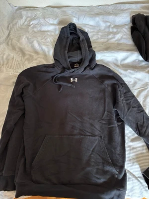 Svart hoodie från Under Armour - Säljer en svart hoodie från Under Armour med klassisk logga på bröstet. Hoodien har dragsko i huvan och en stor magficka framtill. Tillverkad i mjukt och bekvämt material, perfekt för chill eller träning.