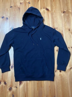 Marinblå hoodie från Polo Ralph Lauren - Snygg marinblå hoodie från Polo Ralph Lauren med klassisk huva och dragsko. Ikoniska vita loggan broderad på bröstet och stor känguruficka framtill. Tillverkad i mjukt bomullsmaterial och har ribbade muddar vid ärmar och nederkant.