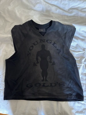 Svart ärmlös topp från YoungLA - Säljer en svart ärmlös topp från YoungLA med tryck av en bodybuilder och texten 'YOUNGLA GOLD'S' på framsidan. Toppen har rund halsringning och är perfekt för gymmet eller träning. Materialet känns mjukt och bekvämt mot huden.