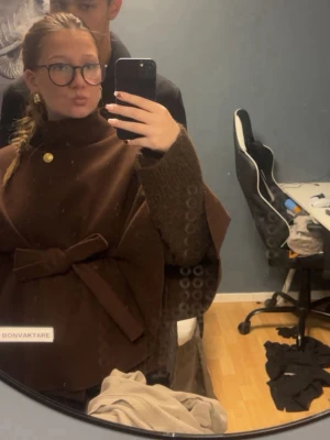 Brun poncho med knytband och guldknapp - Snygg brun ponch med hög krage, dekorativ guldknapp och brett knytband i midjan. Capen har en elegant och lös passform som ger en trendig siluett. Perfekt för dig som vill ha en stilren och unik look. Finns även i vitt, rött och svart.