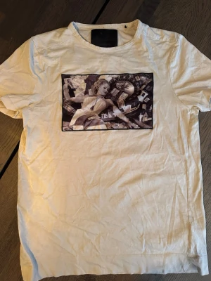 Limitato T-shirt  - Vit t-shirt från Limitato med ett stort svartvitt tryck på bröstet som föreställer en kvinna, pengar och ett roulettebord. T-shirten har rund hals och korta ärmar. Perfekt för dig som gillar unika och uttrycksfulla plagg.