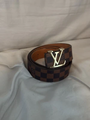 Lv bälte - Snyggt bälte med klassiskt rutigt mönster i brunt och mörkbrunt samt en stor guldfärgad LV-logga som spänne. Perfekt accessoar för att lyfta din outfit och ge en exklusiv touch. Tillverkat i skinnliknande material.