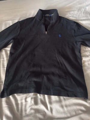Svart halvzip tröja från Polo Ralph Lauren - Klassisk svart långärmad tröja från Polo Ralph Lauren med halv dragkedja och blå broderad logga på bröstet. Tröjan har hög krage och är gjord i mjukt bomullsmaterial. Perfekt för en stilren och avslappnad look.