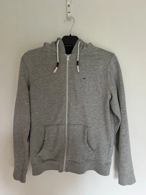 Grå zip hoodie från Tommy Hilfiger - Pris kan minskas vid snabb affärSnygg grå hoodie från Tommy Hilfiger Denim. Zip-up modell med huva, vita dragskor med marinblå/röda detaljer och klassisk Tommy Hilfiger-logga på bröstet. ✨ Stilren och lättmatchad grå färg ✨ Full zip ✨ Huva med dragsko ✨ Två framfickor ✨ Ribbstickade muddar och nederkant ✨ Tommy Hilfiger Denim-detaljer  Storlek: M Färg: Grå / melerad Märke: Tommy Hilfiger Denim  Bra skick enligt bilder – se gärna bilderna för detaljer. Perfekt till jeans, chinos eller mjukisbyxor
