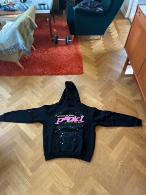 Svart Sp5der Web Hoodie – XL - Svart hoodie med stort spindelnätsmotiv, rosa text och färgglada stjärndetaljer framtill och på ärmarna. Mjuk insida, huva och känguruficka. Helt oanvänd och i nyskick. Se bilder för detaljer.