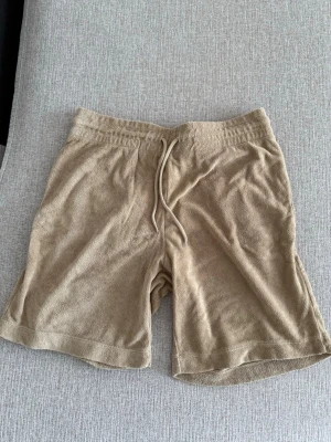 Beige frottéshorts med snörning - Mysiga beige shorts i mjuk frotté med elastisk midja och snörning framtill. Shortsen har en bakficka och avslappnad passform, perfekta för varma dagar eller chill hemma.