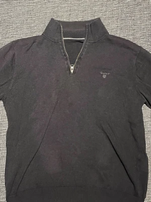 Svart halvzip tröja från GANT - Klassisk svart tröja från GANT med halv dragkedja och ståkrage. Diskret GANT-logga broderad på bröstet. Tröjan har ribbade muddar vid ärmslut och nederkant, perfekt för en stilren och avslappnad look.