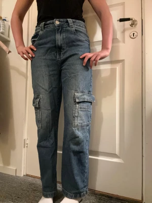 Blå cargobyxor i denim - Säljer ett par blå cargobyxor i klassisk denim med raka ben och stora fickor på sidorna. Byxorna har normal midja, bälteshällor och två bakfickor. Perfekta för en avslappnad och trendig streetwear-look.