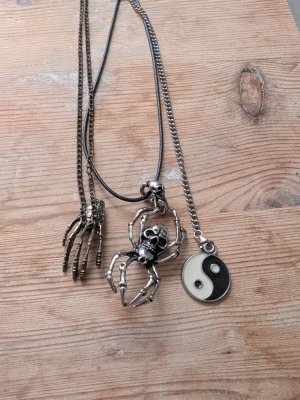 Set med tre halsband med coola hängen/gothic  - Säljer ett set med tre halsband: ett med ett silverfärgat skelett-handhänge, storlek över 30 cm, ett med en stor spindel med dödskalle i metall i storlek 30 längst ut på spännet och ett med ett yin-yang-hänge i svart och vitt, 24 cm. Kedjorna är i metall och läderimitation. Perfekt för dig som gillar alternativ stil och unika detaljer.