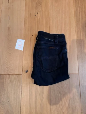 Svarta jeans från Nudie Jeans - Snygga svarta jeans från Nudie Jeans med klassisk femficksmodell och kontrastsömmar på bakfickan. Jeansen har normal midja och raka ben, perfekta för en clean och stilren look. Storlek w29 L32