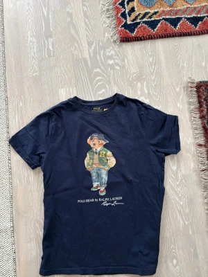 Mörkblå Polo Bear t-shirt Ralph Lauren - Säljer en mörkblå t-shirt från Ralph Lauren med den ikoniska Polo Bear-trycket på bröstet. T-shirten har rund hals och korta ärmar. Perfekt för dig som gillar klassisk streetwear med en twist.